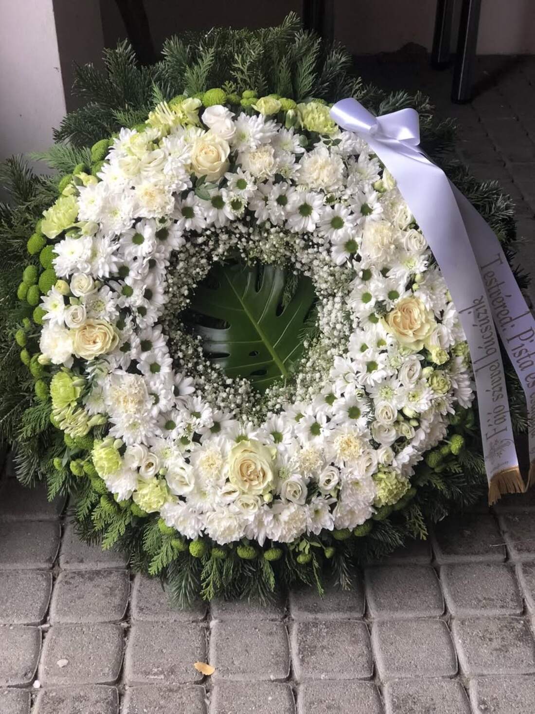 Kerek temetési koszorú fehér krizantémból és rózsából, fenyőalapon, fehér szalaggal.